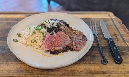 A plate of sliced Dijon Mustard & Paprika-Crusted Standing Rib Roast showcasing the juicy, tender meat with a crispy, flavorful crust. The plate also includes rice and black beans garnished with cheese and scallions.